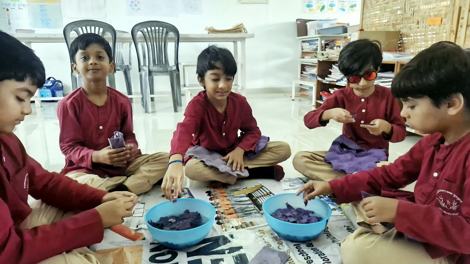 Four children in maroon sit on the classroom floor, each sorting small objects into a blue bowl