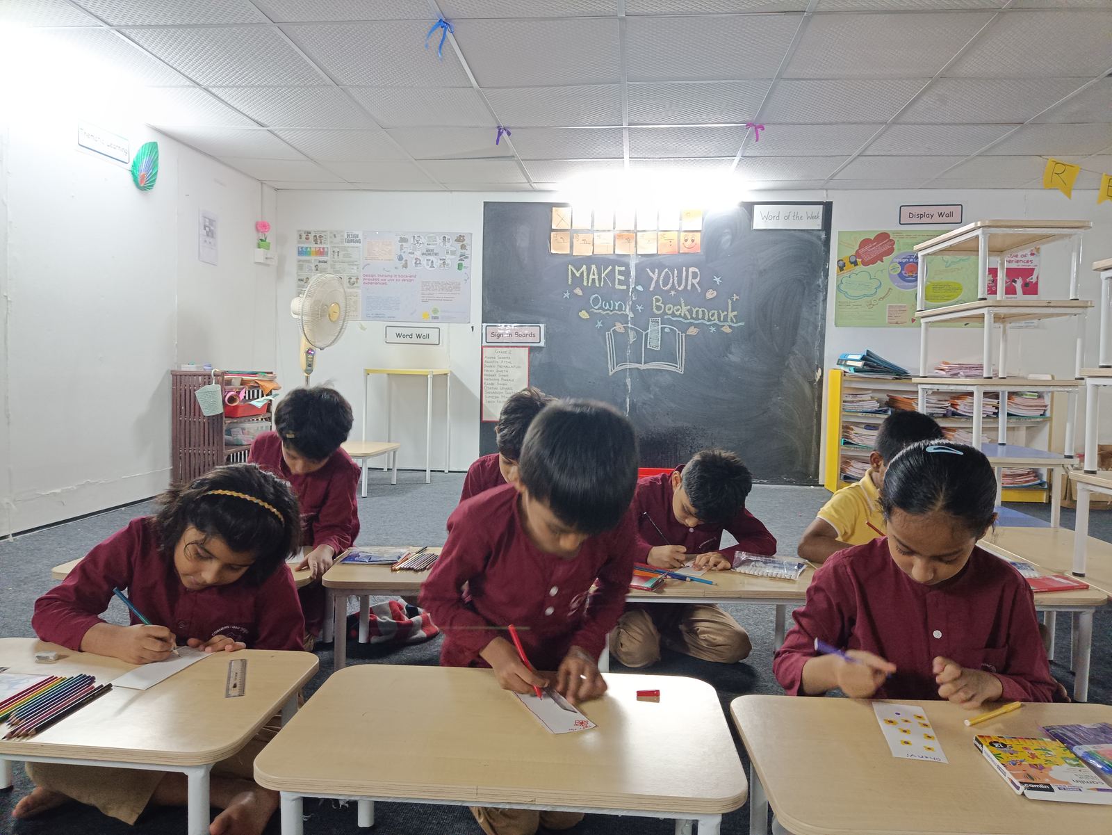 Children in maroon kurtas bend over their desks in a Gurukul classroom, drawing and writing