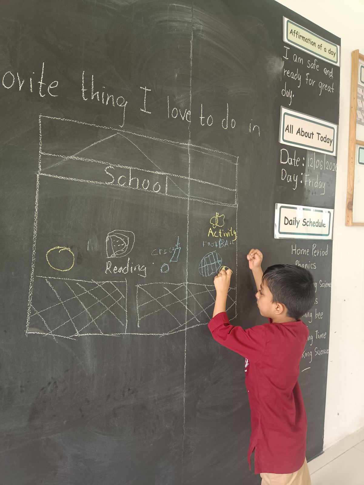 A child in maroon draws on a chalkboard, a sketch of the school beside them