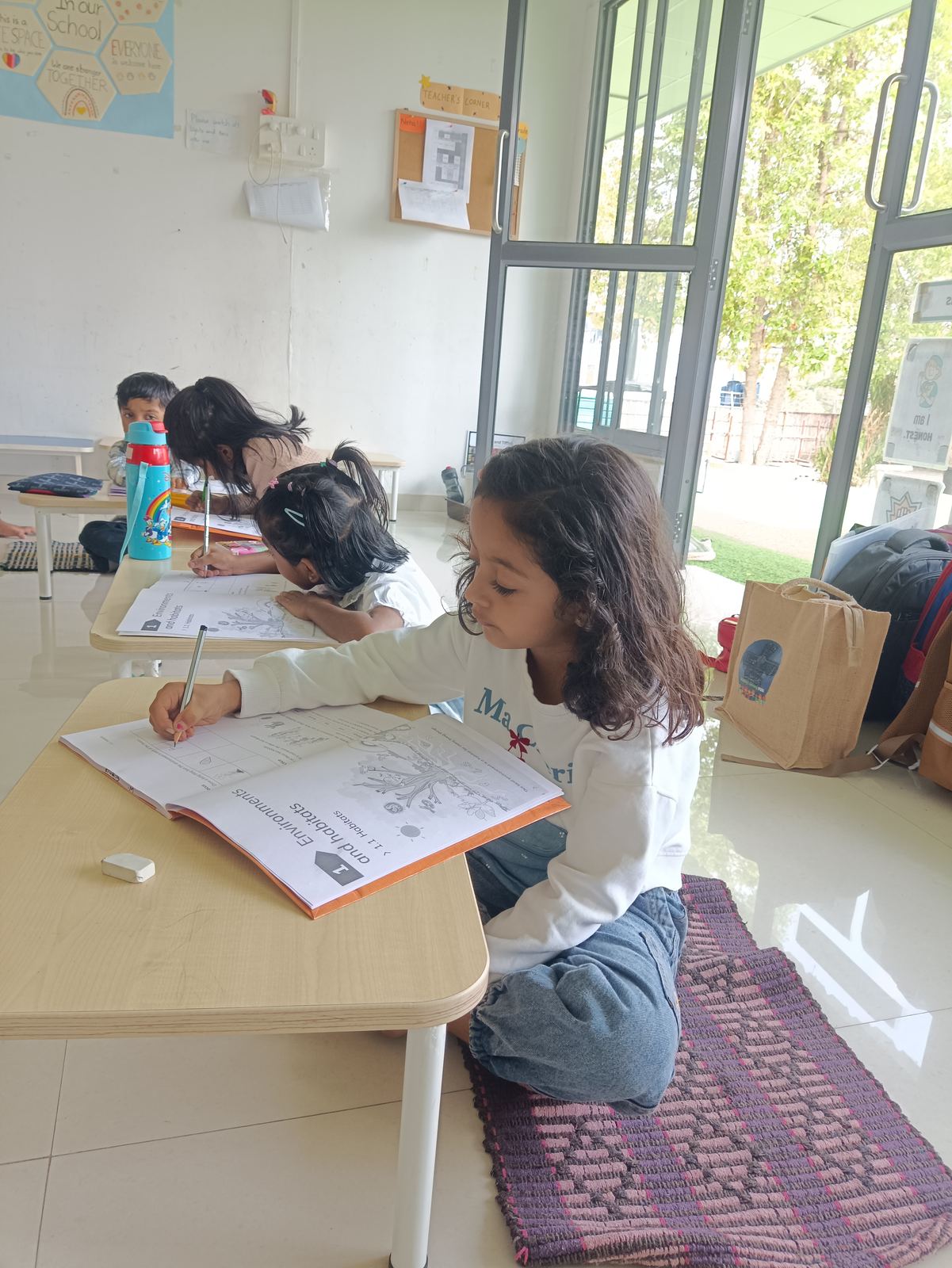 A girl in a white top writes in her workbook at a low desk, in a bright classroom