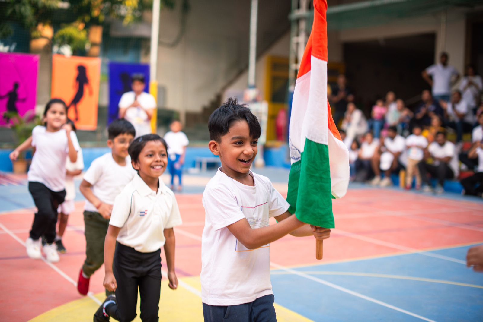A smiling boy in a white T-shirt runs across a painted court carrying an Indian flag, with three classmates running behind him and an audience watching