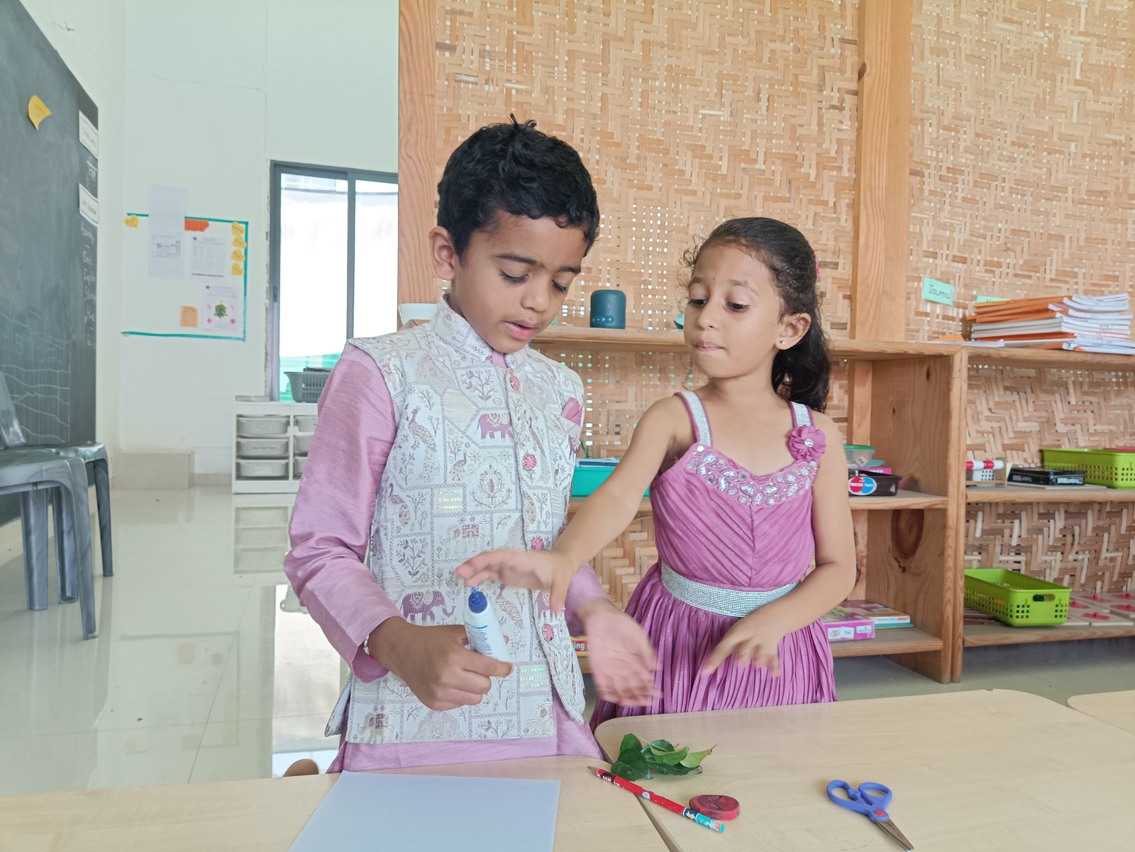 A boy and a girl in festive clothes glue leaves onto a card at a desk