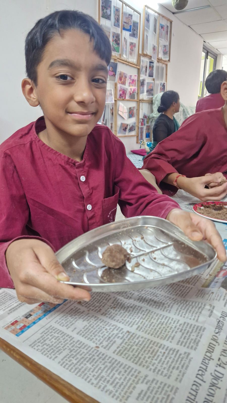 A boy in a maroon kurta smiles and holds up a steel tray with a freshly rolled laddoo