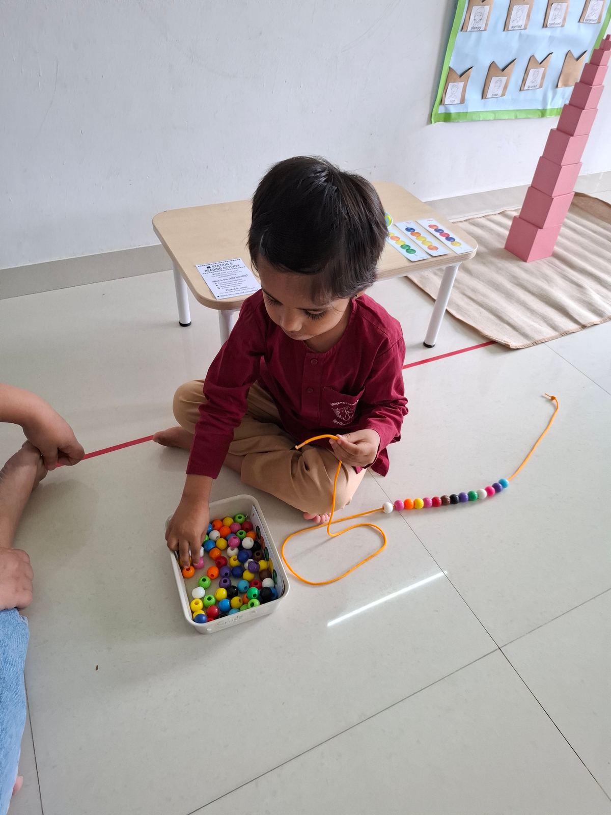 A small boy in maroon sits on the floor and sorts coloured beads from a tray