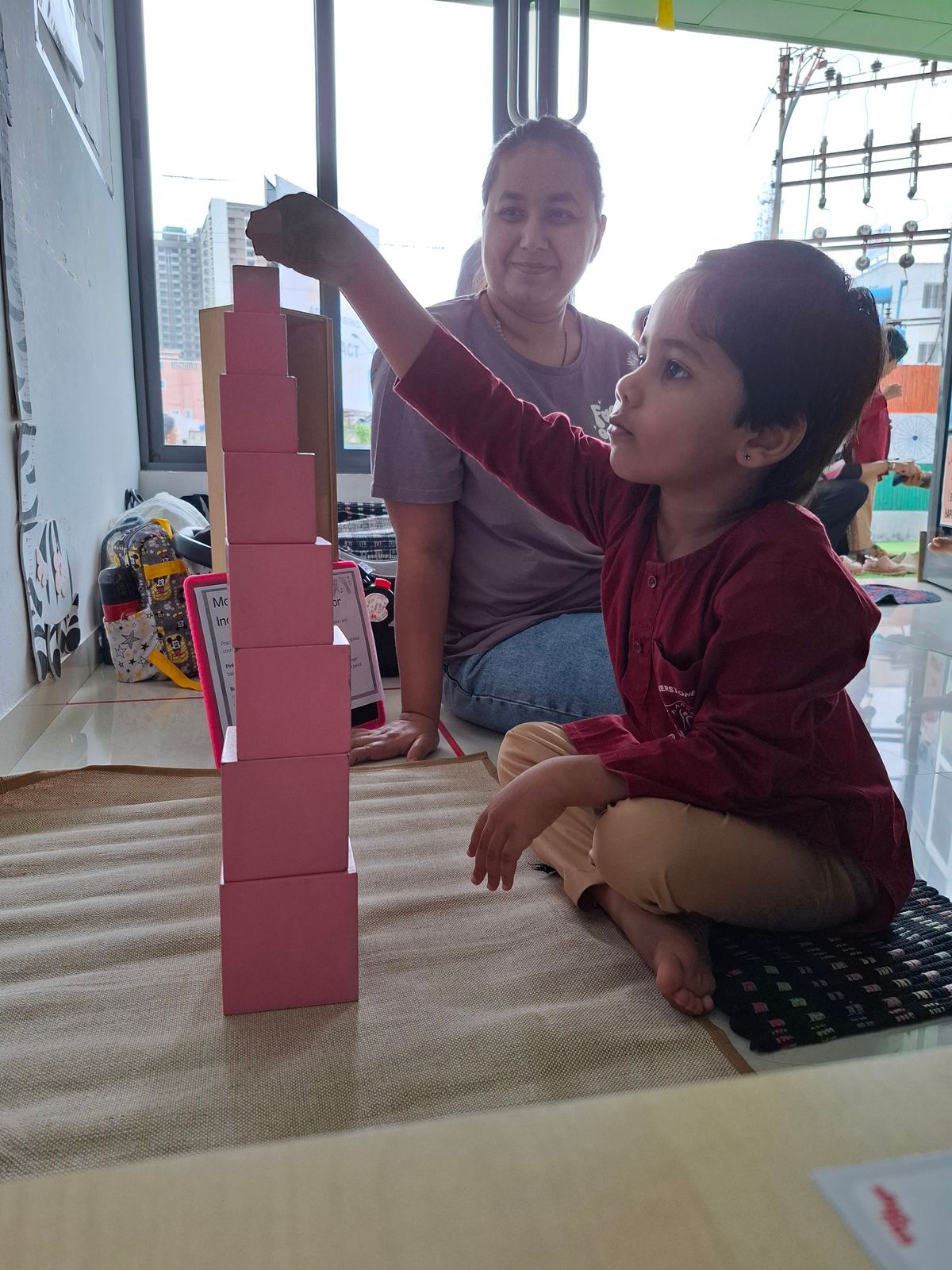 A girl in maroon builds with the pink tower while a teacher looks on