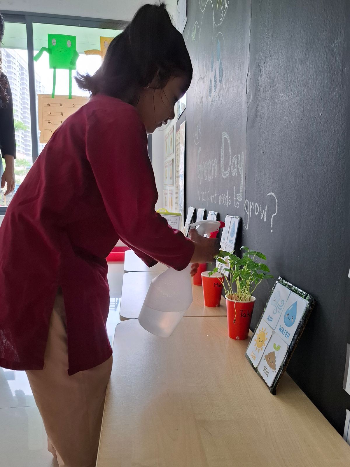 A girl in a maroon kurta tends potted plants on a shelf beside a chalkboard