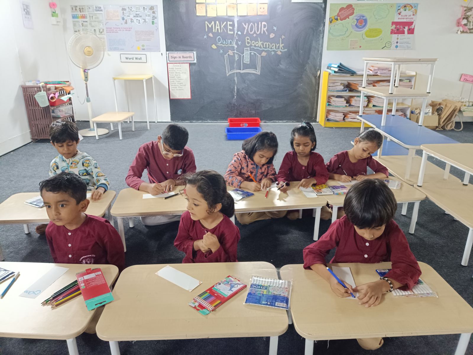 Children in maroon kurtas draw at low desks in a bright classroom