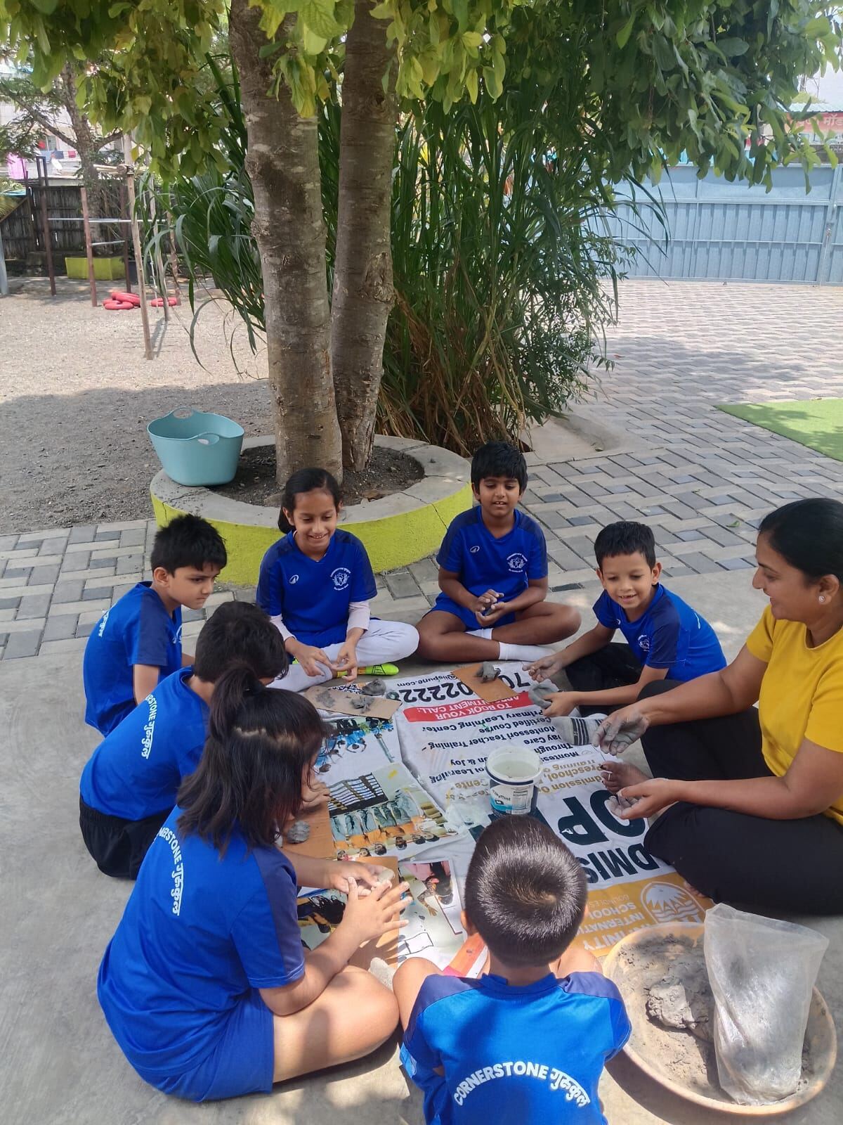 A teacher and children sit in a circle under a tree, working with clay