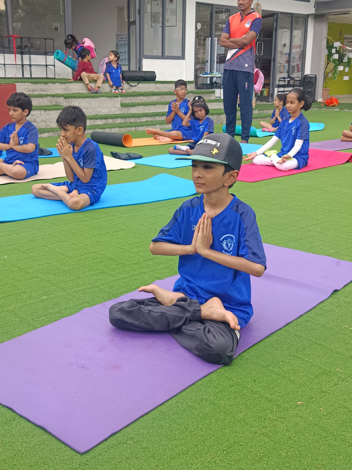 A boy in blue kit sits in lotus pose with his hands together on a mat on the turf