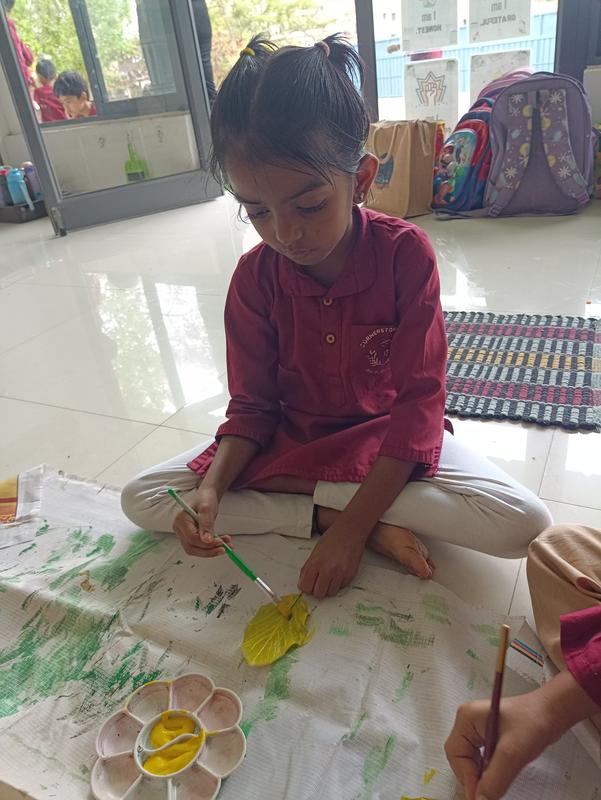 A girl in a maroon kurta sits cross-legged and paints a yellow leaf on large paper